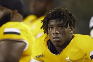 A football player wearing a yellow jersey with the text "CUSA," looking focused while on the sidelines.