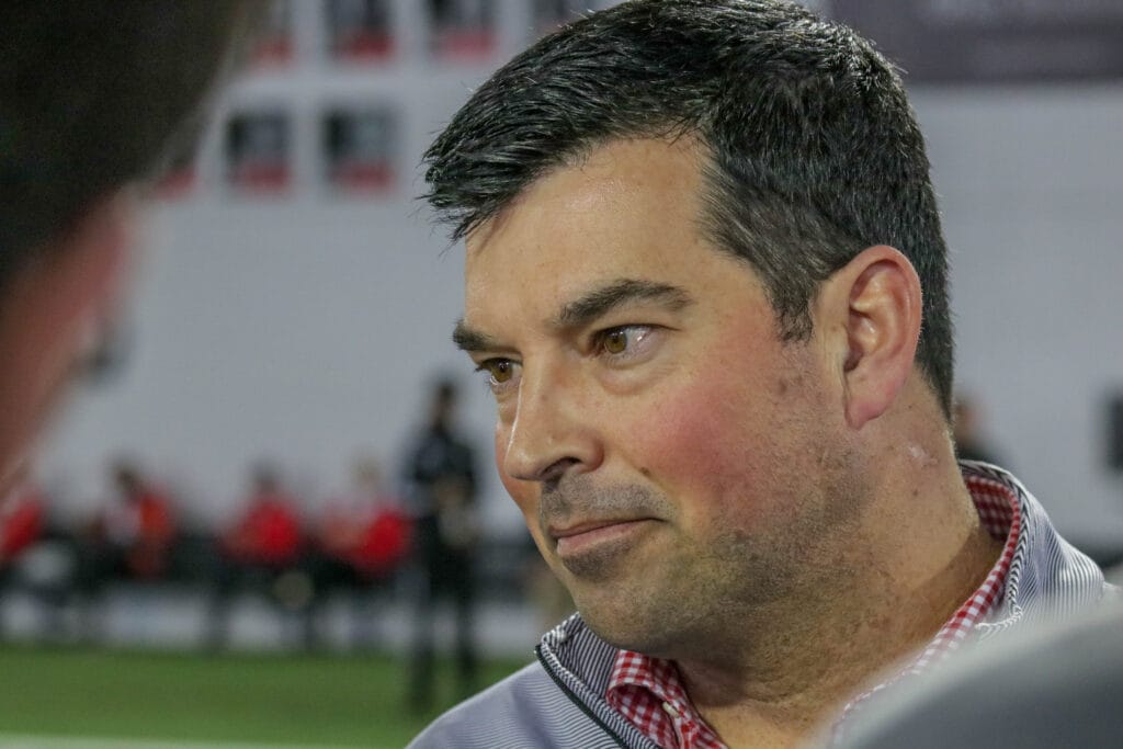 A man in a gray jacket and red checkered shirt looks to the side indoors, exuding the steady focus of a coaching staff member.