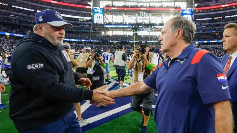 Two football coaches shake hands on the field after a game, surrounded by photographers and staff.